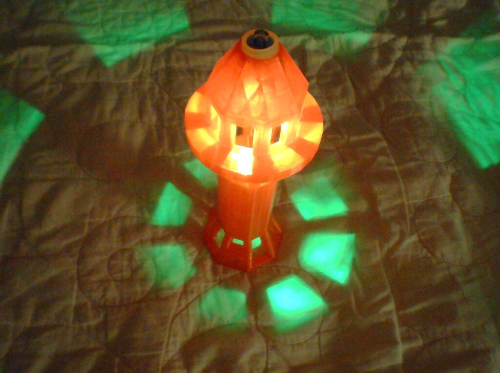 Colorful lantern casting green and orange patterns on a quilted surface. The glowing shape adds a cozy atmosphere, enhancing the room's decor.